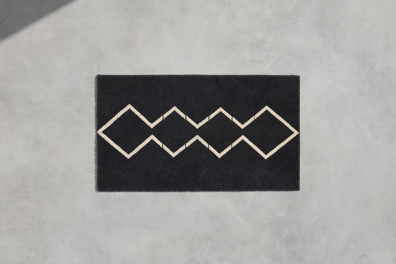 May include: A rectangular black doormat featuring a centered, continuous geometric pattern of four connected diamond shapes outlined in a light beige color. The mat has a textured, fibrous surface and a clean, straight-edged rectangular border.