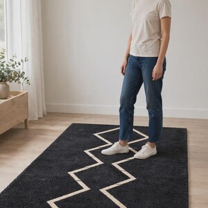 May include: A rectangular area rug featuring a deep charcoal gray shaggy texture with a prominent, contrasting white geometric zigzag line pattern running through the center. The rug is positioned flat on a light-colored wooden floor surface.