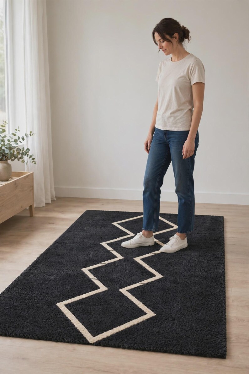 May include: A rectangular area rug featuring a deep charcoal gray shaggy texture with a prominent, contrasting white geometric zigzag line pattern running through the center. The rug is positioned flat on a light-colored wooden floor surface.