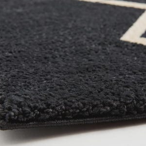 May include: A thick, dark charcoal gray rug with a high-pile, textured surface. The rug features a clean, straight-edged border and a prominent, sharp-angled white geometric line pattern that cuts across the dark, plush fabric surface.
