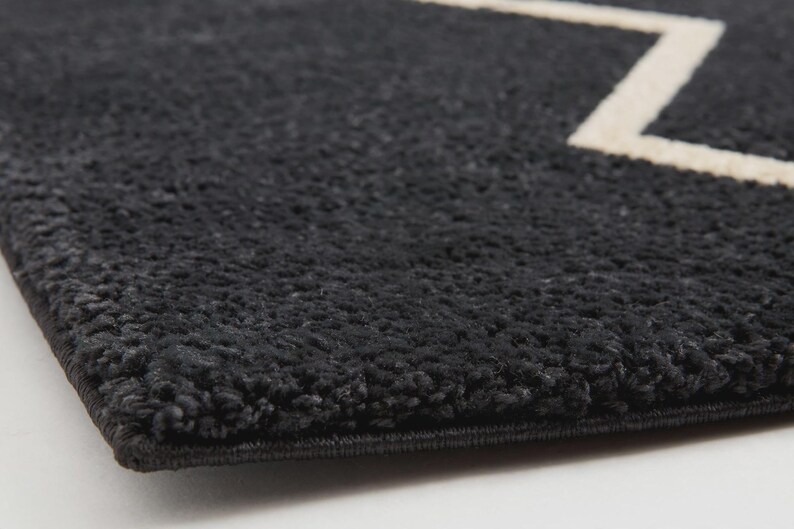 May include: A thick, dark charcoal gray rug with a high-pile, textured surface. The rug features a clean, straight-edged border and a prominent, sharp-angled white geometric line pattern that cuts across the dark, plush fabric surface.