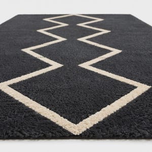May include: A rectangular area rug featuring a deep charcoal gray, high-pile textured surface. A prominent, continuous cream-colored line forms a sharp, symmetrical zigzag pattern that runs vertically through the center of the rug.