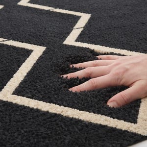 May include: A thick, dark charcoal gray rug featuring a prominent, light cream-colored zigzag pattern. The rug has a deep, plush, high-pile texture that shows visible indentations when pressed, highlighting the soft and dense construction of the material.