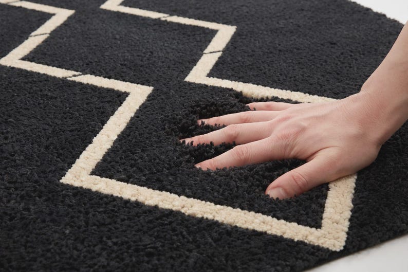 May include: A thick, dark charcoal gray rug featuring a prominent, light cream-colored zigzag pattern. The rug has a deep, plush, high-pile texture that shows visible indentations when pressed, highlighting the soft and dense construction of the material.