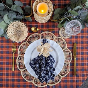 Peut inclure: Mise en place avec une assiette blanche, une serviette à motifs bleu marine et un anneau de serviette en forme de fleur tissée. Des couverts dorés et un dessous de plat tissé sont disposés sur une nappe à carreaux. Une bougie allumée et des éléments décoratifs complètent la scène.