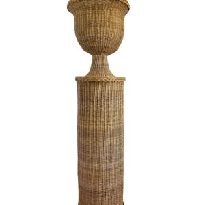 May include: A tall, woven wicker planter with a pedestal base. The planter has a large, urn-shaped top and a cylindrical base. The wicker is a natural tan color, and the piece has a classic, elegant design.