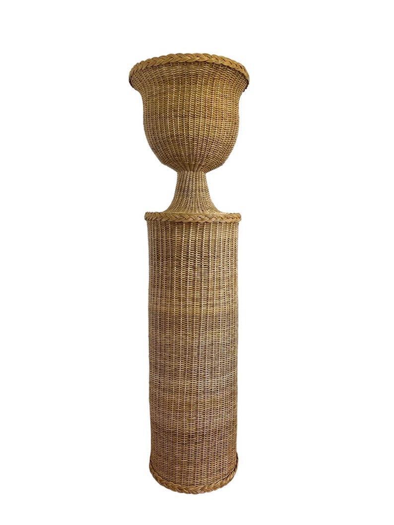 May include: A tall, woven wicker planter with a pedestal base. The planter has a large, urn-shaped top and a cylindrical base. The wicker is a natural tan color, and the piece has a classic, elegant design.