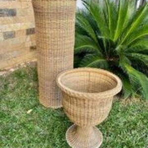 May include: Two wicker planters, one tall and cylindrical, the other a pedestal planter with a rounded bowl. Both are woven in a natural tan color, set on green grass with a palm tree in the background. The cylindrical planter is approximately 36 inches tall.