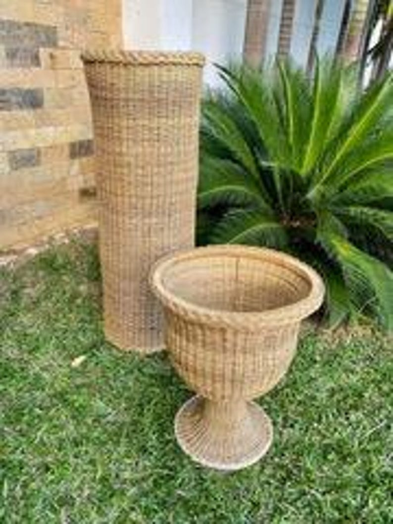 May include: Two wicker planters, one tall and cylindrical, the other a pedestal planter with a rounded bowl. Both are woven in a natural tan color, set on green grass with a palm tree in the background. The cylindrical planter is approximately 36 inches tall.