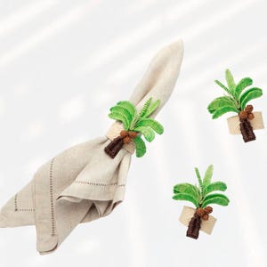 May include: A beige linen napkin is held by a green and brown palm tree napkin ring. Two additional palm tree napkin rings are also visible. The palm trees have green fronds, brown trunks, and beige bases.