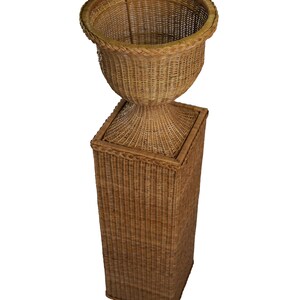 May include: A tall, woven wicker planter with a pedestal base. The planter has a wide, open top and a rounded body. The base is a square column. The wicker is a natural, light brown color. The planter is suitable for indoor or outdoor use.