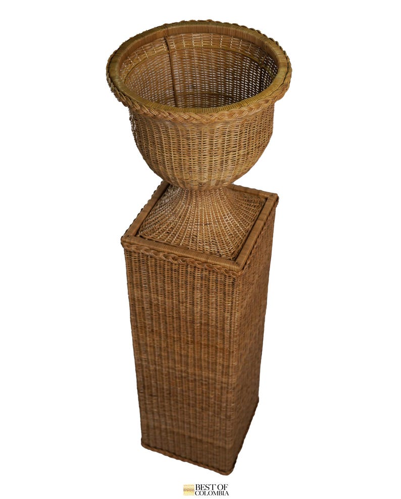 May include: A tall, woven wicker planter with a pedestal base. The planter has a wide, open top and a rounded body. The base is a square column. The wicker is a natural, light brown color. The planter is suitable for indoor or outdoor use.