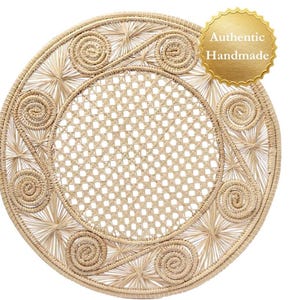 Puede incluir: Mantel individual circular tejido en tono beige claro. El diseño incluye espirales y estrellas alrededor del borde, y un centro tejido. Una pegatina dorada en la esquina superior derecha dice "Auténtico Hecho a Mano".
