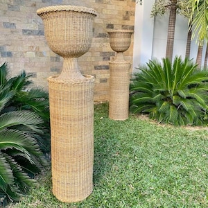 May include: Two woven wicker planters with a natural finish. The planters have a pedestal base and a large, bowl-shaped top. One planter is taller than the other. They are set on a grassy lawn with a stone wall in the background.
