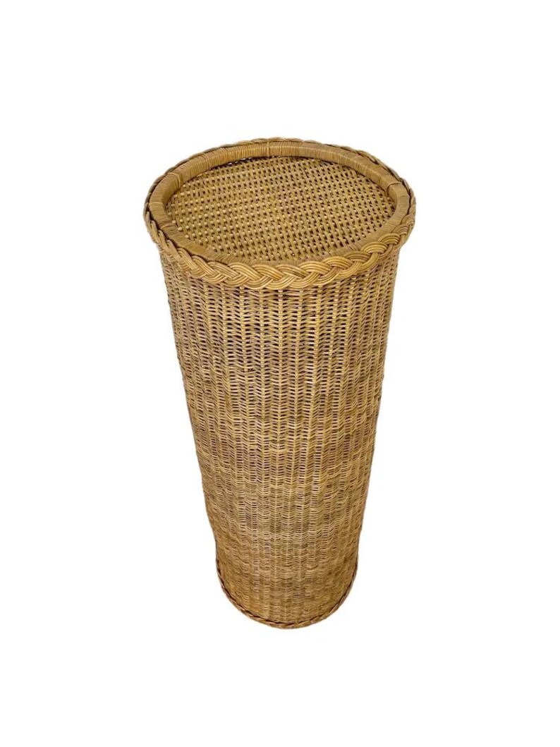 May include: A tall, cylindrical wicker basket with a woven lid. The basket is made of light brown wicker and has a braided trim around the top. The basket is empty and stands on a white background.