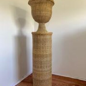 May include: A tall, woven wicker plant stand with a cylindrical base and a decorative, urn-shaped top. The stand is light brown and features vertical ribbing on the base and a flared rim on the top. It is placed in a corner against a white wall.