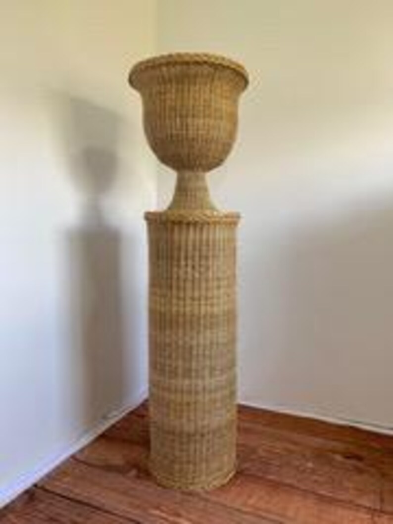 May include: A tall, woven wicker plant stand with a cylindrical base and a decorative, urn-shaped top. The stand is light brown and features vertical ribbing on the base and a flared rim on the top. It is placed in a corner against a white wall.