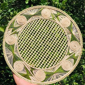 Könnte beinhalten: Runder gewebter Platzdeckchen mit einem Gittermuster in der Mitte und dekorativen Spiral- und Sternmuster in Beige und Grün. Der Platzdeckchen besteht aus Naturfasern und ist vor einem Hintergrund aus grünem Laub angeordnet.