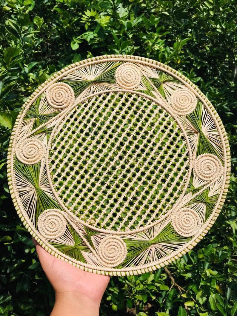 Pode incluir: Sousplat redondo tecido com um centro de treli&ccedil;a e padr&otilde;es decorativos de espirais e explos&otilde;es em bege e verde. O sousplat &eacute; feito de fibras naturais e &eacute; colocado sobre um fundo de folhagem verde.