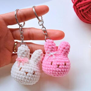 Puede incluir: Dos llaveros de conejito de ganchillo, uno blanco y otro rosa, cada uno con un pequeño lazo y detalles bordados. Los llaveros están sujetos a llaveros y cadenas plateadas. Una madeja de hilo rojo es visible en el fondo.