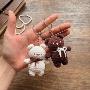 May include: Two handmade crochet bear keychains. One is white with a brown scarf, the other is brown with a white bow. Both have silver key rings attached. The bears are small, cute, and likely soft to the touch.