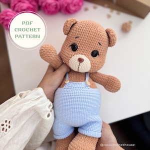 Puede incluir: Un oso de peluche tejido a ganchillo de color marrón que lleva un peto azul claro con botones pequeños. El oso tiene ojos de seguridad negros, un hocico color crema y una nariz marrón. Un gráfico circular blanco a la izquierda dice PDF CROCHET PATTERN en verde.