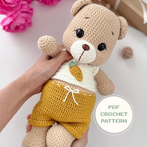 Puede incluir: Un oso de peluche tejido a ganchillo con hilo marrón claro, con ojos de seguridad negros, nariz marrón y hocico blanco. El oso lleva una camiseta blanca sin mangas con un pequeño aplique de zanahoria naranja y pantalones cortos color mostaza.