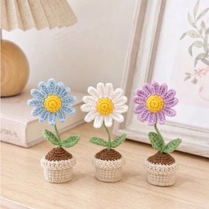 May include: Three crocheted daisy flowers in small pots. The flowers are blue, white, and purple, each with a yellow center. The pots are beige and brown, and the leaves are green. The flowers are displayed on a wooden surface.