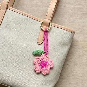 Puede incluir: Un bolso tote de lona beige con asas y ribetes de cuero marrón claro. Un charm de flor de ganchillo rosa y magenta con una hoja verde cuelga del asa. El charm está sujeto con un cordón rosa y un anillo dorado.