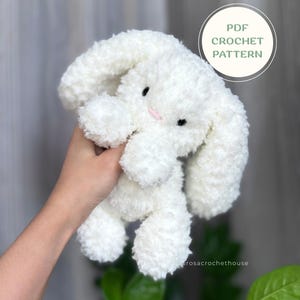 Op de afbeelding: Een wit pluche konijn-knuffel met lange slappe oren, zwarte veiligheidsoogjes en een kleine roze geborduurde neus. Een cirkelvormige afbeelding rechtsboven bevat de tekst PDF CROCHET PATTERN. De tekst rosacrochethouse is ook zichtbaar.