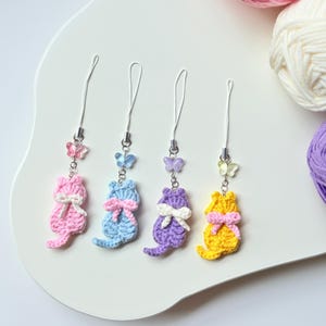 Puede incluir: Cuatro colgantes de gato de ganchillo en rosa, azul, morado y amarillo, cada uno con un lazo a juego y un charm de mariposa. Los colgantes están sujetos a cordones blancos. Se ve lana de varios colores en el fondo.