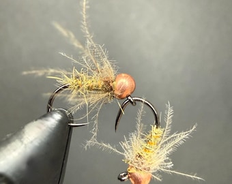 May include: Two artificial fishing flies featuring tan, fuzzy, synthetic fibers and a metallic copper-colored bead head. Each fly is constructed on a curved, dark-colored metal hook, designed to mimic aquatic insect larvae for fly fishing.