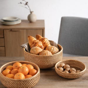 May include: Three woven rattan baskets on a wooden table. One basket holds oranges, another contains bread, and a third holds walnuts. A vase with a sprig of greenery and a stack of plates are in the background.