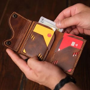 Può includere: Portafoglio in pelle marrone con più fessure per carte e uno scomparto per contanti. Il portafoglio è aperto, rivelando una carta di credito gialla, una carta di credito rossa e una pila di banconote. Il portafoglio è tenuto aperto da mani.