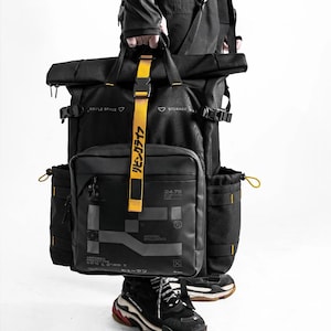 Puede incluir: Mochila negra con cierre enrollable, múltiples bolsillos y detalles amarillos. La bolsa presenta un bolsillo cuadrado frontal con detalles gráficos y texto japonés en una correa amarilla. La bolsa es sostenida por una persona.