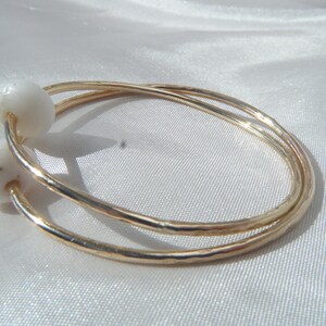 May include: Gold-toned bangle bracelets with white, oval-shaped accents. The bracelets are stacked and have a hammered texture. The jewelry is displayed on a white, satin-like surface.