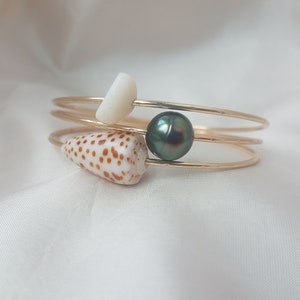 May include: A gold bangle bracelet with three strands, adorned with a white shell, a brown and white seashell, and a dark green pearl.
