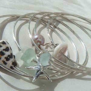 May include: A set of six silver bangles with various charms, including a seashell, a starfish, a pearl, and a button. The charms are arranged around the bangles, creating a layered and whimsical look.