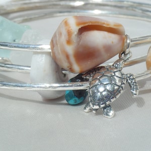Può includere: Un set di quattro braccialetti d'argento con una varietà di ciondoli, tra cui una conchiglia, una perla bianca, una perla turchese e un ciondolo a forma di tartaruga d'argento.