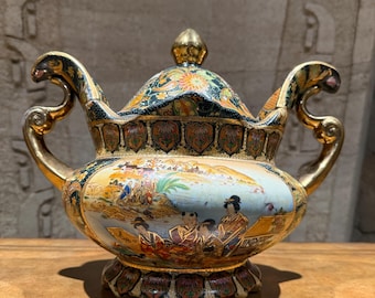 May include: An ornate, covered ceramic vessel with gold accents and handles. The vessel features a detailed painted scene with figures in traditional clothing, set against a landscape. The lid is topped with a gold finial, and the overall design is highly decorative.