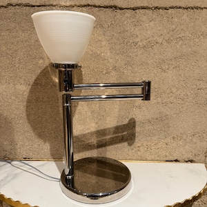 May include: A chrome-finished desk lamp with a white, cone-shaped shade. The lamp features an adjustable arm and a round base. The shade has a textured, ribbed design. The lamp is approximately 15 inches tall.