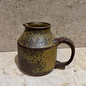 May include: A small, brown and green ceramic pitcher with a speckled glaze. The pitcher has a rounded body and a handle.