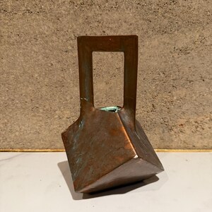 May include: A copper-colored, geometric vase with a rectangular handle. The vase has a unique, angular design and a weathered patina, suggesting age. The vase is displayed on a white surface against a textured, neutral-toned background.
