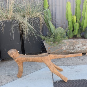 May include: A rustic wooden stool with a unique, human-like design. The stool is made of natural wood and has a rough, textured surface. It is sitting on a concrete surface.