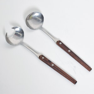 May include: A set of two stainless steel serving utensils with wooden handles. The utensils have a polished finish and feature a spoon and a slotted spoon.