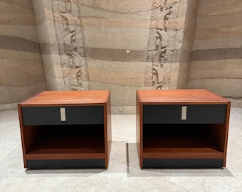 May include: Two matching wooden nightstands with a mid-century modern design. Each nightstand features a reddish-brown wood finish, a black drawer with a silver-colored handle, and a black base. The nightstands are placed on a light-colored floor against a textured wall.