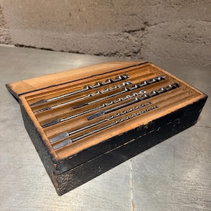 May include: A set of five metal auger drill bits arranged in a rectangular wooden storage box. The box features a natural wood interior with grooved channels for each bit and a dark, weathered exterior finish with a hinged lid design.