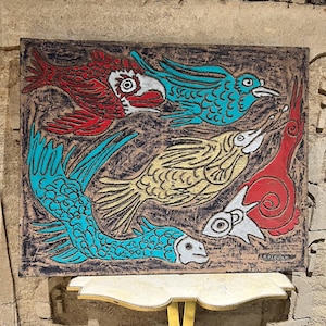 May include: A textured, rectangular artwork featuring stylized birds and fish in red, teal, white, and yellow against a brown background. The artwork has a rustic, hand-crafted appearance, with visible brushstrokes and a signature in the lower right corner.