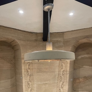 May include: A white ceiling light fixture with a round, flat shade hangs from a white ceiling with a black beam. The light is on and casting a warm glow.