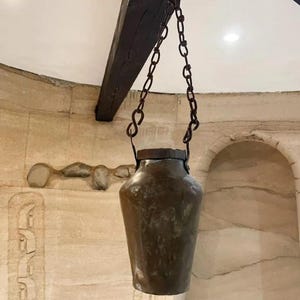 May include: An antique copper hanging pot with a chain and a lid. The pot is hanging from a wooden beam in a room with a stone wall.